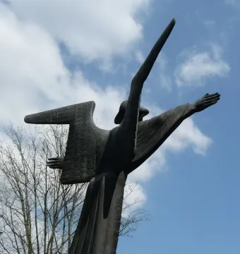 The Angel of Peace sculpture shown from below against a blue sky with clouds, with one arm extended and a pointed form rising beside it.