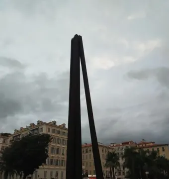 Neuf Lignes Obliques stands in a paved square with palm trees, buildings, and people nearby.