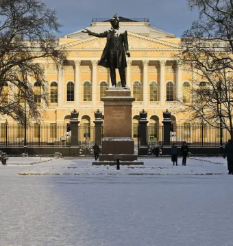 Pushkin statue on a pedestal in a snowy park, with a yellow building and bare winter trees in the background.