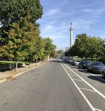 Trenton Battle Monument rises in the distance beside an American flag along a tree-lined street.
