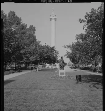 Washington Monument rises above a park with trees, lawns, paths, and a seated statue in the foreground.