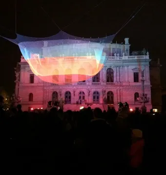 1.26 illuminated mesh sculpture glowing in bands of orange, yellow, and blue above a crowd near the Rudolfinum at night.
