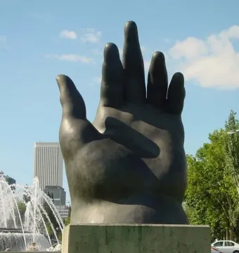 Botero’s Hand sculpture on a pedestal in a Madrid park with a fountain and buildings behind it.