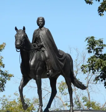 Elizabeth II equestrian statue silhouetted against a clear blue sky, framed by green trees.