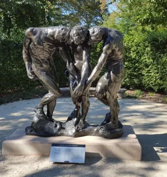 The Three Shades by Auguste Rodin, a dark bronze sculpture of three intertwined male figures with bowed heads outdoors near greenery.