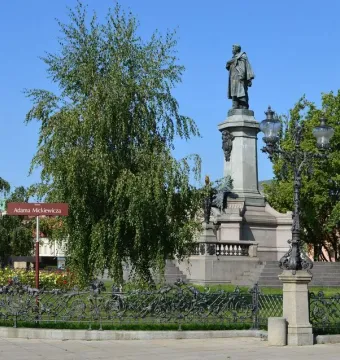 Adam Mickiewicz Monument with a bronze cloaked statue on a stone pedestal in Warsaw, framed by tree leaves and rose bushes.