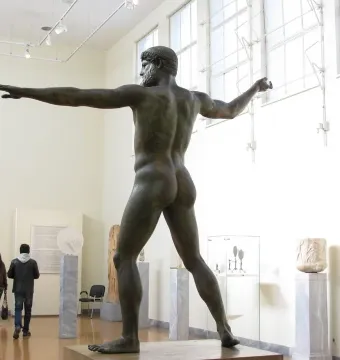 Artemision Bronze shown from the back and left side in a museum gallery, with one arm extended and the other raised.