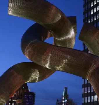 A golden sculpture of intertwined metallic tubes stands against a twilight sky with city buildings in the background.