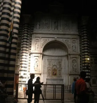 Piccolomini Altarpiece inside a dimly lit church with black-and-white striped columns and visitors nearby.