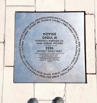Commemorative plaque for the Sea Organ embedded in light-colored paving stones, with hiking shoes visible at the bottom.