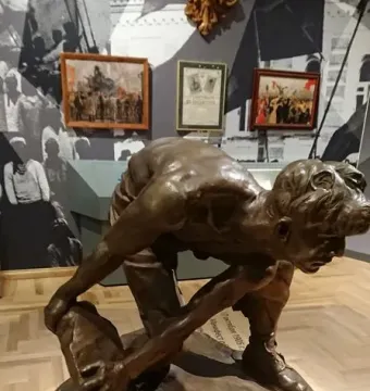 Bronze sculpture of a muscular man bent over gripping a large stone, displayed on a pedestal in a museum.