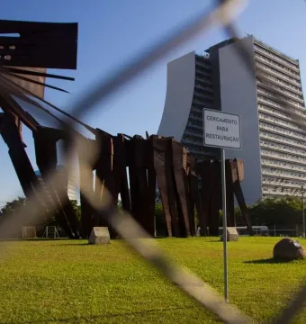 Monument to the Azoreans seen behind a chain-link fence, with a modern building and a restoration sign in the background.