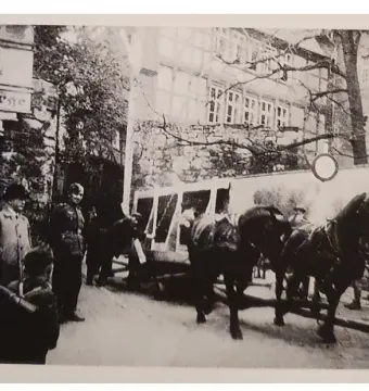 Horse-drawn covered vehicle moving through a street with several people and buildings in the background.