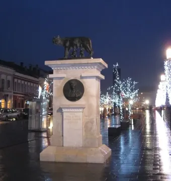 The Capitoline Wolf sculpture on a tall stone pedestal along a wet street at twilight, with historic buildings and lit lampposts nearby.