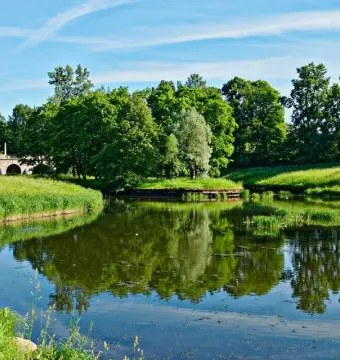 A slender monument to Elena Pavlovna stands near a multi-arched stone bridge over calm water surrounded by green trees.
