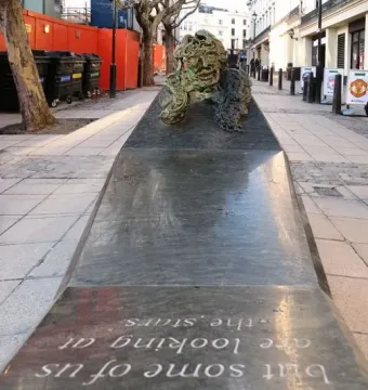 A long dark stone monument with an oxidized bronze bust and engraved text, set on a paved street with buildings and trees behind it.