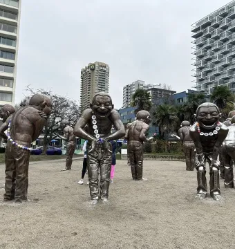 A-maze-ing Laughter statues standing on sandy ground with city buildings and trees behind them.