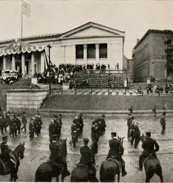 A large parade of uniformed men marches along a wet street past a decorated building, with the Charles Devens statue covered by an American flag and crowds gathered nearby.