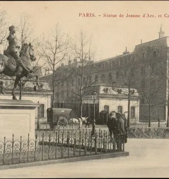 Equestrian statue of Joan of Arc on a tall pedestal behind an iron fence, with a large building and bare trees in the background.