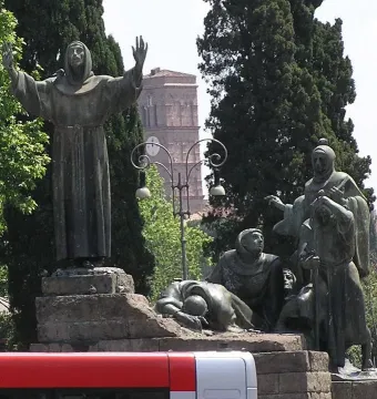 Bronze monument to Saint Francis of Assisi with a hooded figure standing above kneeling figures beside a stone base.