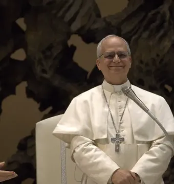 An elderly cleric in white vestments with glasses and a crucifix necklace smiles beside a microphone, with another cleric partly visible at left.
