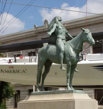Appeal to the Great Spirit bronze statue of a Native American rider on horseback with arms outstretched, set against a sailboat and stone building.