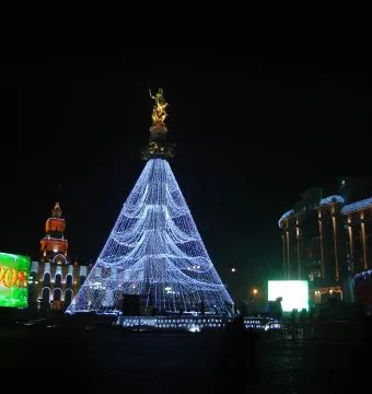 The Freedom Monument topped with a gilded figure, surrounded by bright blue lights forming a towering cone at night.