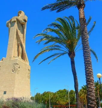 Monument to Columbus seen from below, framed by palm trees against a blue sky.