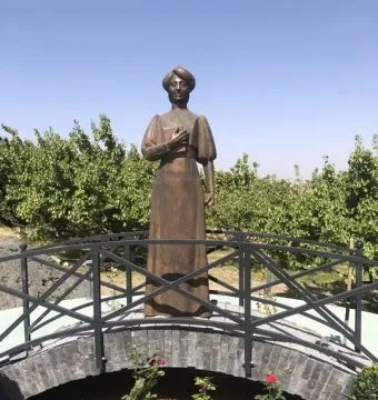 Bronze statue of Zabel Yessayan holding a book, standing on an arched bridge with roses and trees behind her.