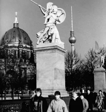 Athena Protects the Young Hero statue on a pedestal with pedestrians nearby, with the Berlin Cathedral and TV tower in the background.