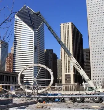 Cloud Gate’s segmented metal shell under construction with a crane overhead and Chicago buildings in the background.