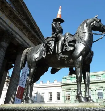 Duke of Wellington bronze equestrian statue with a traffic cone on the rider’s hat, set against a grand building under a blue sky.