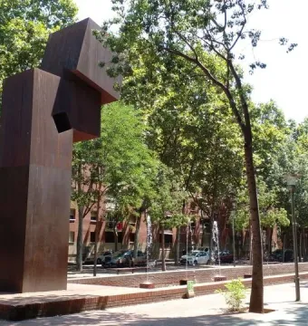 El Llarg Viatge, a tall dark abstract sculpture with a square opening beside a fountain and trees.