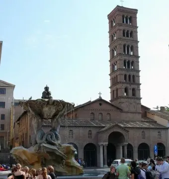 Fountain of the Tritons with sculpted figures supporting a shell-like basin, water cascading into the basin below, with a church campanile in the background.