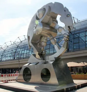 Rolling Horse sculpture beside a modern station with a vaulted glass roof, shown on a wedge-shaped pedestal with round porthole windows.