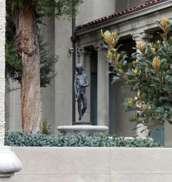 Spirit of Spanish Music bronze statue of a boy holding a long pipe, framed by foliage and a courtyard building.