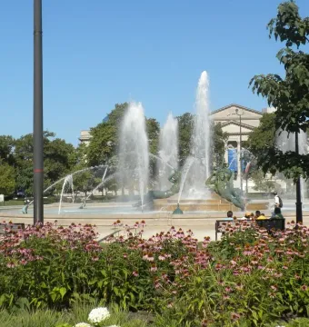 Swann Memorial Fountain rises behind a patch of pink flowers, with water shooting high from the circular basin.