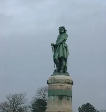 The Vercingetorix Monument, a bronze statue on a tall stone pedestal, stands on a grassy hill beneath an overcast sky.