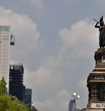 Cuauhtémoc Monument with a bronze figure in a feathered headdress on an ornate pedestal, set against modern buildings and trees.