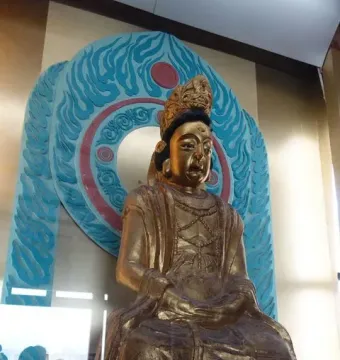 A gilded seated statue with a crown and ornate robes displayed against a carved blue and red backdrop at Huangze Monastery Rock Sculptures.