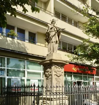Weathered stone statue of John of Nepomuk holding a cross, mounted on an ornate pedestal behind a spiked fence.