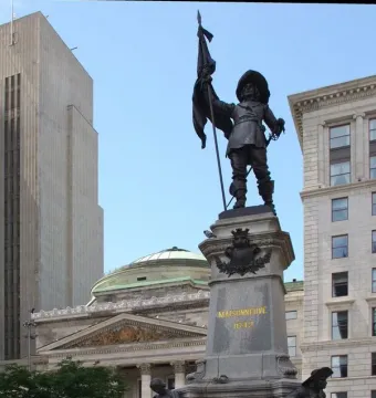 Maisonneuve Monument with a bronze figure holding a flag and sword atop a stone pedestal inscribed “MAISONNEUVE 1642,” with smaller figures below and city buildings in the background.