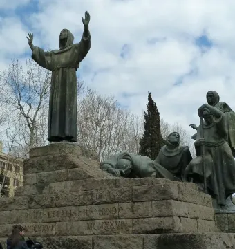 Monument to Saint Francis of Assisi with dark bronze figures on a stone plinth, including a hooded figure raising his hands toward the sky.