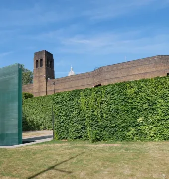 The National Police Memorial stands as a tall translucent green glass monument beside a shallow pool, with an ivy-covered wall and brick building behind it.