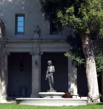 Spirit of Spanish Music bronze statue on a stone fountain basin before a building with pillars and archways.