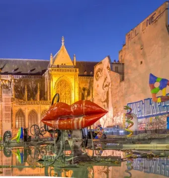Stravinsky Fountain with colorful abstract sculptures, including red lips and a bird-like figure, beside an illuminated Gothic building at dusk.