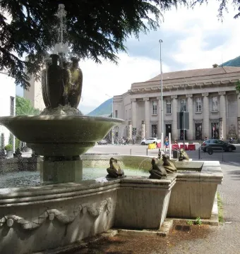 The Fountain of the Frogs stone fountain sprays water in a city square with a large classical building in the background.