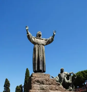 Bronze Monument to Saint Francis of Assisi with robed figures on a stepped stone plinth under a blue sky.