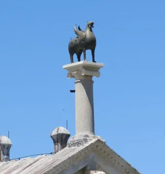 Pisa Griffin statue on a tall stone column above the roof of a cathedral, against a blue sky.
