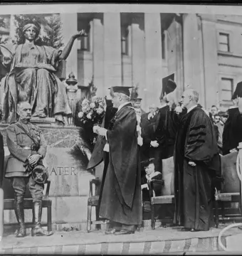 Alma Mater, a bronze seated woman with a laurel wreath, scepter, and open book, on a marble pedestal surrounded by people.